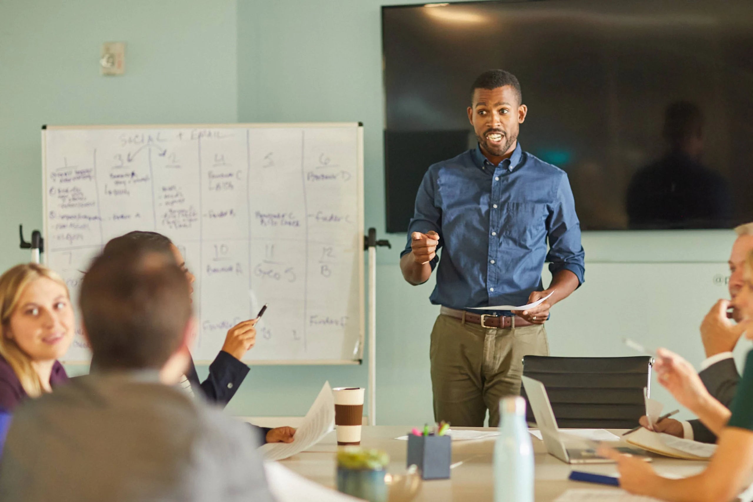 Man standing at a table in a conference room talking with others sitting at the table.
