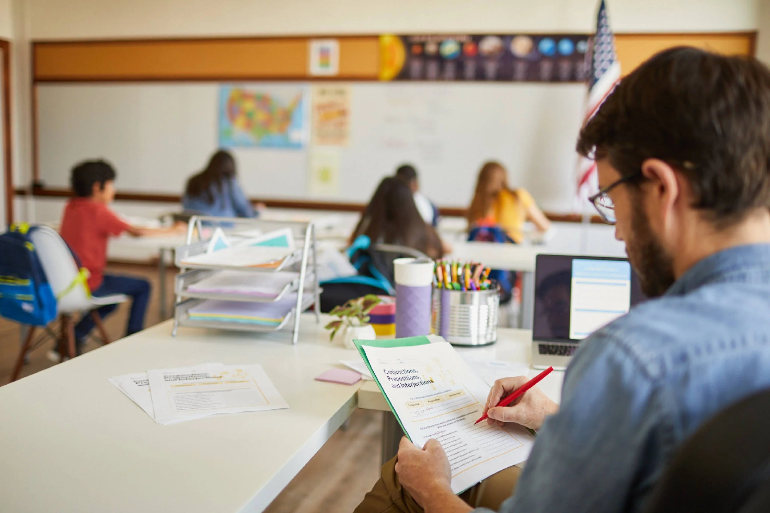 Teacher grading a worksheet with students in the background.